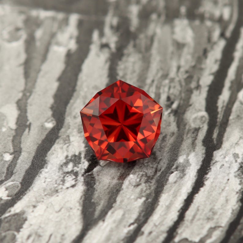 Puede incluir: Una piedra preciosa roja con forma octogonal facetada. La piedra preciosa est&aacute; colocada sobre un fondo gris y blanco.