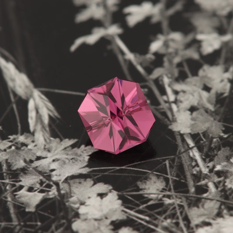 May include: A pink hexagonal gemstone with a star-shaped pattern inside. The gemstone is sitting on a black background with a blurry image of white flowers.