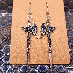 May include: A pair of silver-colored axe-shaped earrings. The earrings are displayed on a brown card against a blue, textured background. The axes have detailed designs on the blades and handles, with a hook at the top for hanging.