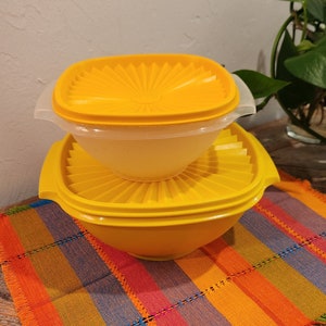 May include: A set of two yellow plastic bowls with lids. The bowls are stacked on top of each other. The bowls have a ribbed design and are sitting on a colorful striped placemat.
