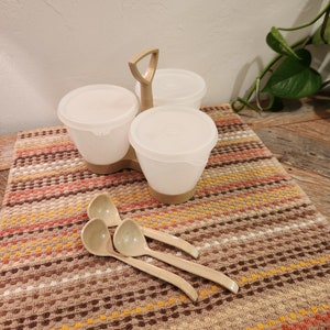 May include: A vintage kitchen set featuring three white plastic containers with lids, connected by a tan handle. Three matching tan serving spoons are arranged in front of the containers. The set rests on a patterned brown and orange cloth.