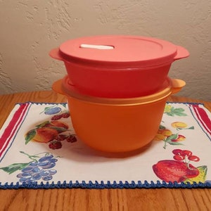 May include: Two plastic storage containers, one orange and one pink, stacked on top of each other. The containers are on a white tablecloth with a red and blue border.
