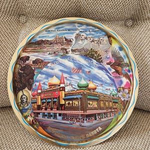 May include: A round metal tray with a colorful design featuring images of Mount Rushmore, Badlands National Monument, a corn palace, and a pheasant. The tray has a blue and gold border and the text "Mitchell, South Dakota" in the bottom center.