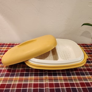 May include: A yellow plastic food steamer with a lid and a white perforated insert. The steamer is on a red and black plaid tablecloth.