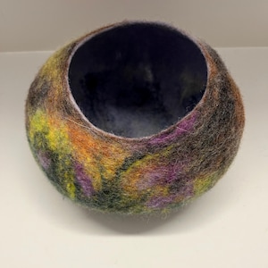 May include: A round, felted bowl featuring a textured surface with a blend of dark purple, orange, yellow, and green fibers. The interior of the bowl is a deep, solid dark purple, creating a contrast with the multi-colored exterior pattern.