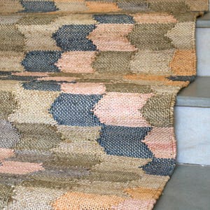 Puede incluir: Una alfombra tejida con un patrón geométrico en tonos marrones, rosas y azules. La alfombra está hecha de fibras naturales y tiene un aspecto rústico y hecho a mano.