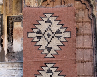May include: A brown and beige patterned rug with a geometric design. The rug has a fringe border and is hanging on a wall.