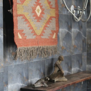 May include: A bathroom scene featuring a decorative rug with a geometric pattern in shades of orange, gray, and beige, hanging on a tiled wall. A wooden shelf holds two small sculptures. The wall is tiled in a dark gray marble pattern.