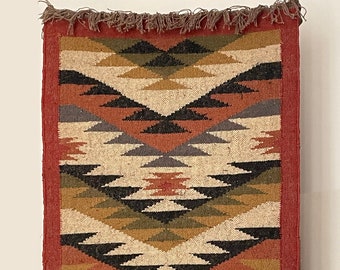 May include: A rectangular, woven wall hanging with a geometric pattern. The design features diamond and triangle shapes in shades of beige, black, brown, green, and orange, framed by a red border. The top edge has a fringe.