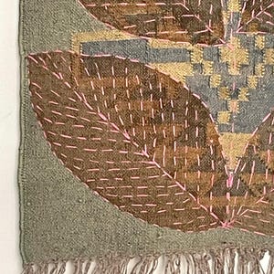May include: Textile wall hanging with a leaf design. The piece features brown, grey, and gold tones, with pink stitching outlining the leaf shapes. The edges are a muted green, and the bottom has a fringe.