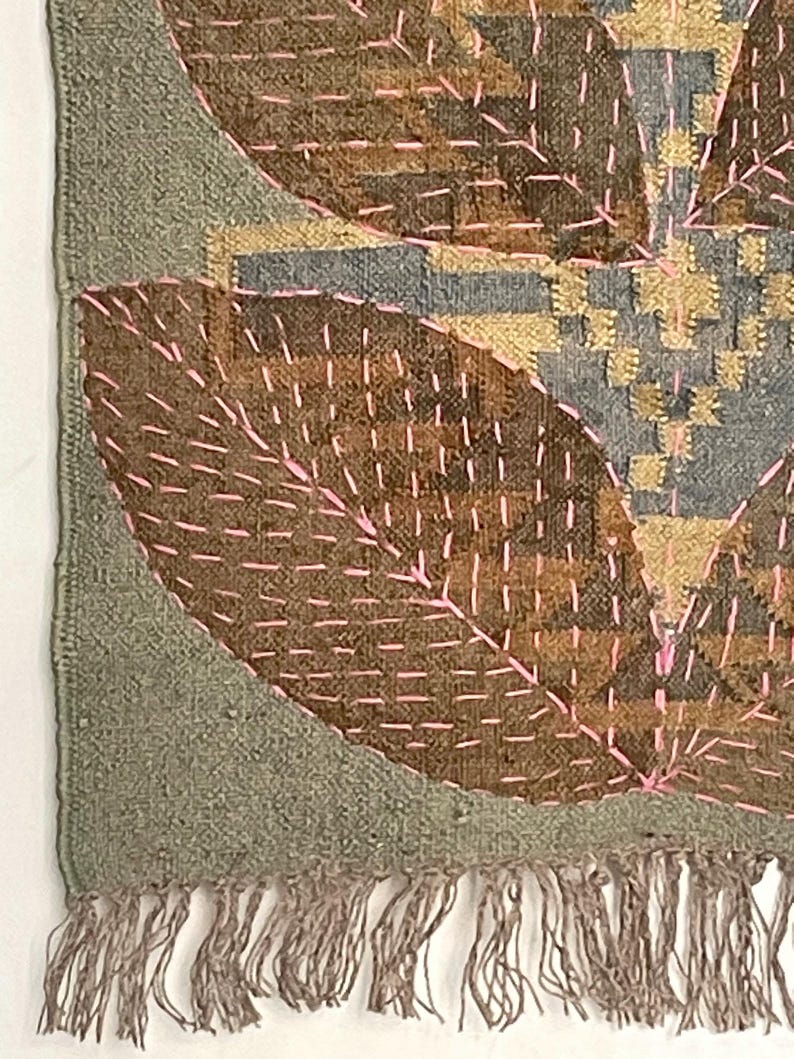 May include: Textile wall hanging with a leaf design. The piece features brown, grey, and gold tones, with pink stitching outlining the leaf shapes. The edges are a muted green, and the bottom has a fringe.