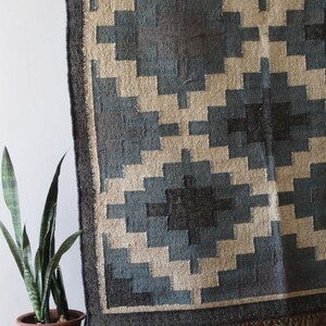 May include: A blue and beige geometric patterned rug with a black border and fringe. The rug is displayed on a wooden stand.