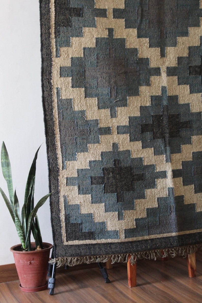 May include: A blue and beige geometric patterned rug with a black border and fringe. The rug is displayed on a wooden stand.