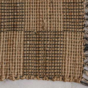 K&ouml;nnte beinhalten: Ein gewebter Teppich in nat&uuml;rlicher brauner Farbe mit schwarzen Streifen. Der Teppich hat einen Fransenrand.
