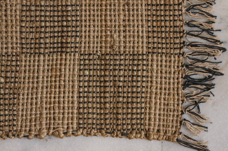 K&ouml;nnte beinhalten: Ein gewebter Teppich in nat&uuml;rlicher brauner Farbe mit schwarzen Streifen. Der Teppich hat einen Fransenrand.