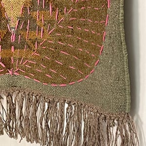May include: Close-up of a textile art piece with a textured, woven appearance. The piece features a combination of olive green, brown, and gold hues, with bright pink stitching. The bottom edge is finished with a fringe of brown threads. The design appears to be abstract.