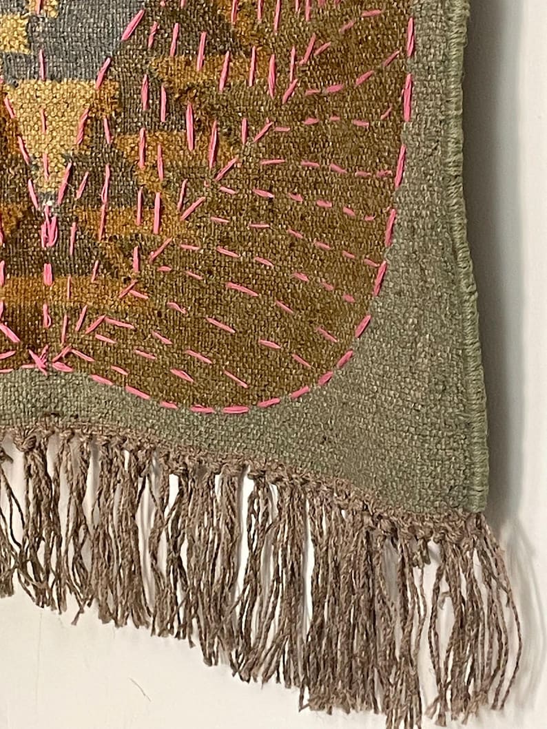 May include: Close-up of a textile art piece with a textured, woven appearance. The piece features a combination of olive green, brown, and gold hues, with bright pink stitching. The bottom edge is finished with a fringe of brown threads. The design appears to be abstract.