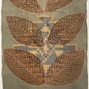 May include: A rectangular textile with a green backdrop and brown, leaf-shaped designs outlined in pink stitching. The centre features geometric patterns in gold and grey, with a fringed edge at the bottom. Likely a wall hanging or decorative piece.