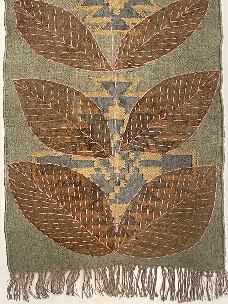 May include: A rectangular textile with a green backdrop and brown, leaf-shaped designs outlined in pink stitching. The centre features geometric patterns in gold and grey, with a fringed edge at the bottom. Likely a wall hanging or decorative piece.