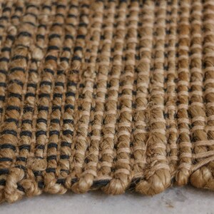 K&ouml;nnte beinhalten: Nahaufnahme eines gewebten Teppichs in nat&uuml;rlichen Braun- und Schwarzt&ouml;nen. Der Teppich besteht aus einem dicken, strukturierten Material und hat eine leicht raue Oberfl&auml;che.