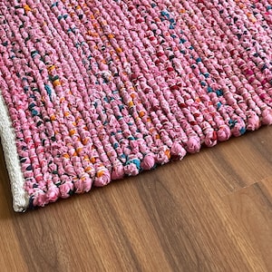 Puede incluir: Una alfombra tejida rosa y blanca hecha de tela reciclada. La alfombra tiene una superficie texturizada y un borde blanco.