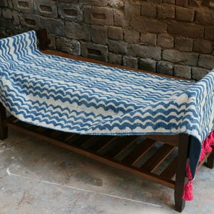 May include: A wooden bench with a blue and white patterned textile draped over it. The textile has a wave-like design and is trimmed with pink tassels. The bench is made of dark wood and sits on a concrete surface.