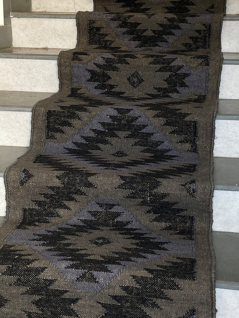 K&ouml;nnte beinhalten: Ein schwarz-grauer Teppich mit geometrischem Muster. Der Teppich ist aus einem gewebten Material gefertigt und hat einen Fransenrand. Der Teppich liegt auf einer Treppe.