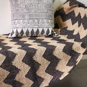 May include: A black and beige woven rug with a chevron pattern. The rug is draped over a wooden bench with a white cushion with a black floral design on top.