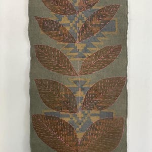 May include: Olive green textile wall hanging with a leaf design. The leaves are brown with pink stitching, and the background features a geometric pattern in grey and gold. The bottom edge has a fringe.