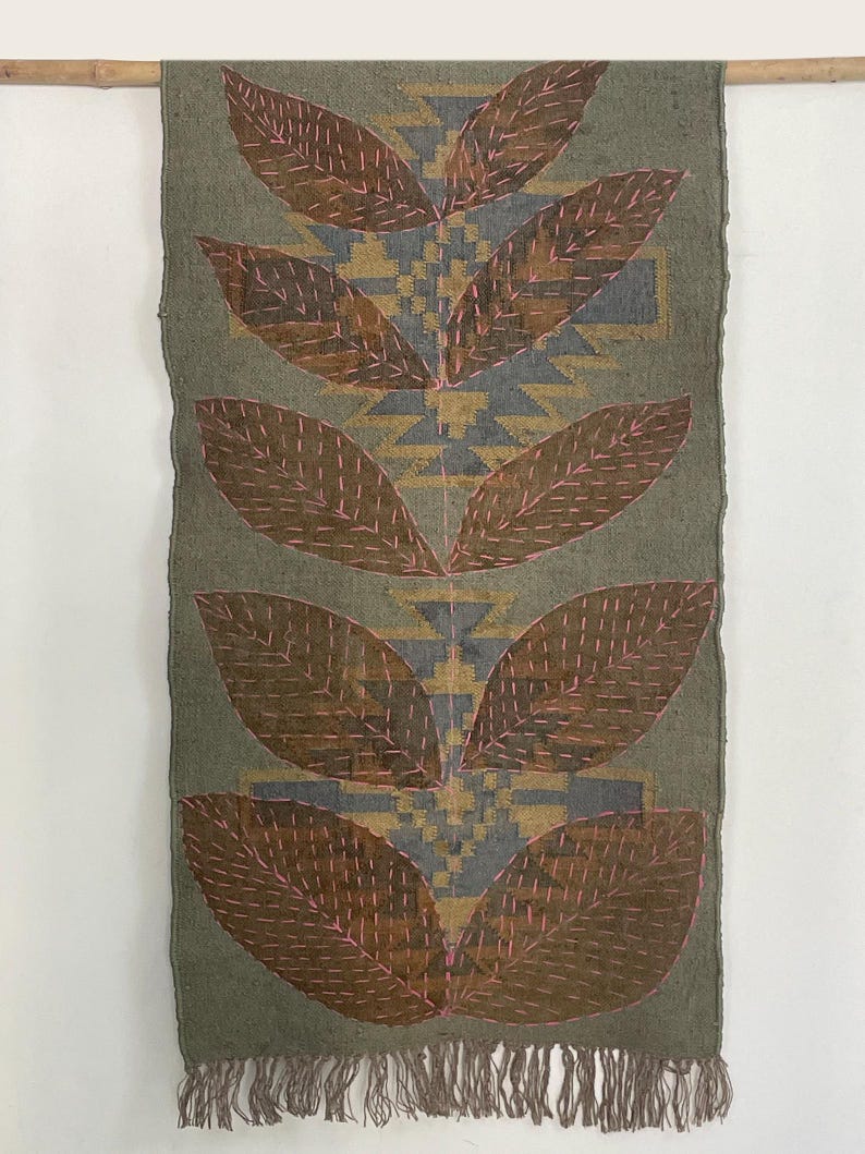 May include: Olive green textile wall hanging with a leaf design. The leaves are brown with pink stitching, and the background features a geometric pattern in grey and gold. The bottom edge has a fringe.