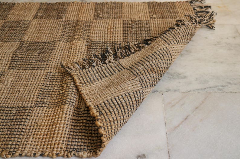 K&ouml;nnte beinhalten: Ein brauner und schwarzer karierter Jute-Teppich mit Fransen an den R&auml;ndern. Der Teppich ist aus Naturfasern gefertigt und hat einen rustikalen Look.