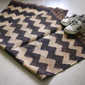 May include: A rectangular rug with a zig-zag pattern in dark brown and beige. The rug has a fringed edge and is next to a pair of white trainers with black stripes.