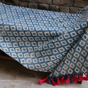 Peut inclure: Une couverture en coton bleu et blanc &agrave; motifs avec des glands rouges. La couverture est pli&eacute;e et drap&eacute;e sur un banc en bois.