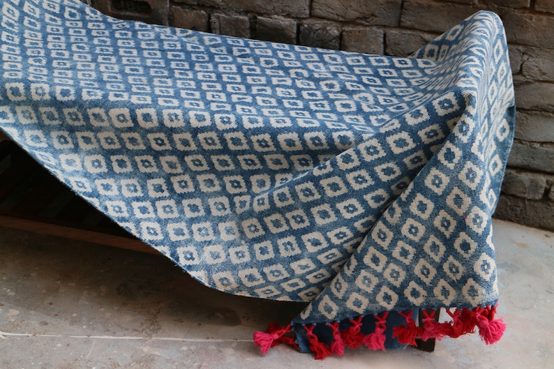 Peut inclure: Une couverture en coton bleu et blanc &agrave; motifs avec des glands rouges. La couverture est pli&eacute;e et drap&eacute;e sur un banc en bois.