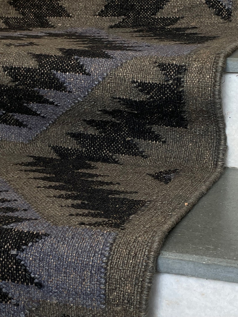K&ouml;nnte beinhalten: Ein grauer und schwarzer Teppich mit geometrischem Muster. Der Teppich ist aus gewebtem Stoff und hat einen Fransenrand.