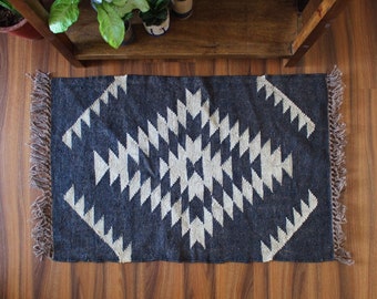 May include: A rectangular, dark blue rug with a geometric diamond pattern in off-white. The rug has a fringed edge in a neutral color. The rug is placed on a wooden floor.