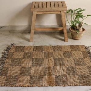 K&ouml;nnte beinhalten: Ein brauner und schwarzer Schachbrettmuster-Jute-Teppich mit Fransen an den R&auml;ndern. Der Teppich liegt auf einem wei&szlig;en Fliesenboden vor einem Holzhocker.