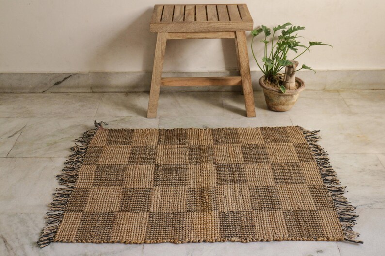 K&ouml;nnte beinhalten: Ein brauner und schwarzer Schachbrettmuster-Jute-Teppich mit Fransen an den R&auml;ndern. Der Teppich liegt auf einem wei&szlig;en Fliesenboden vor einem Holzhocker.