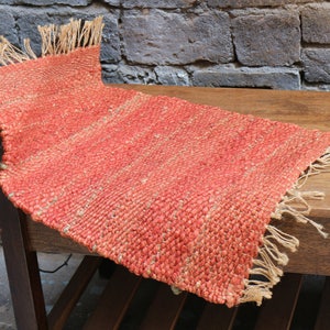 May include: A woven orange and beige rug with fringe on all sides. The rug is laying on a wooden table.