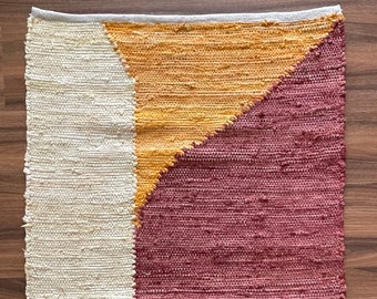 May include: A rectangular woven rug with a geometric design. The rug features three distinct sections: a cream-colored vertical stripe on the left, a golden-yellow triangle in the center, and a burgundy triangle on the right. The rug is made of a textured material.