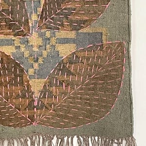 May include: Textile art with a leaf design. It features brown, grey, and gold colours with pink stitching. The bottom edge has a fringe.