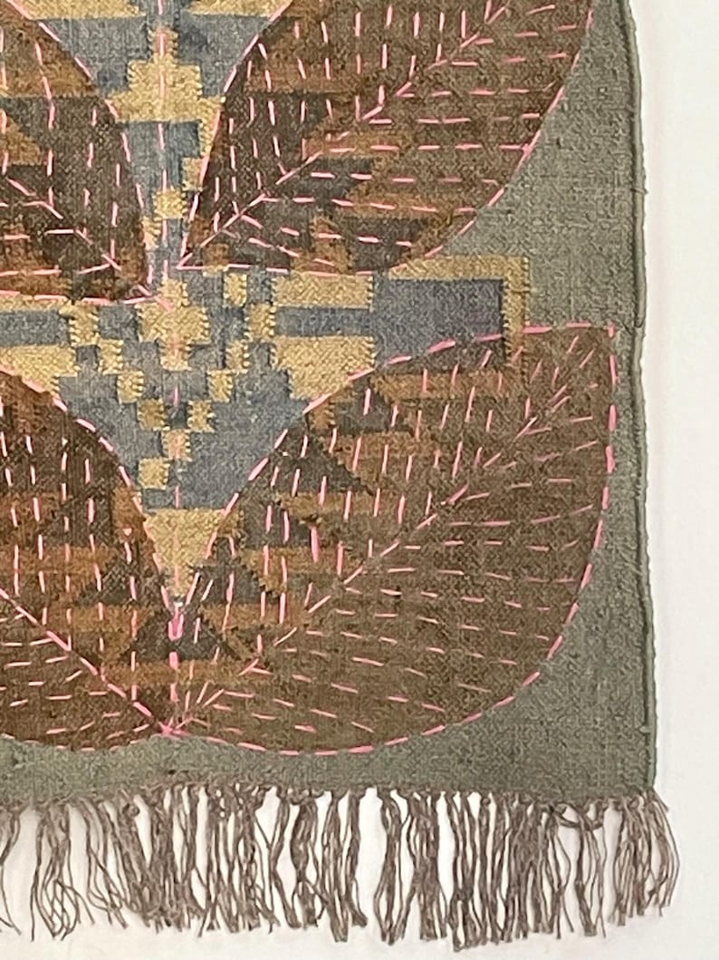 May include: Textile art with a leaf design. It features brown, grey, and gold colours with pink stitching. The bottom edge has a fringe.