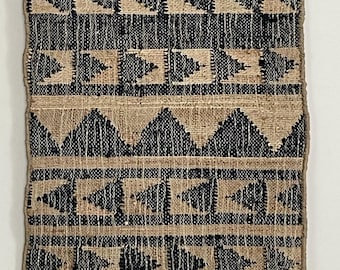 May include: A rectangular wall hanging with a natural jute background and a repeating pattern of black geometric shapes. The design includes triangles, squares, and tree-like figures. The bottom edge has a fringe.