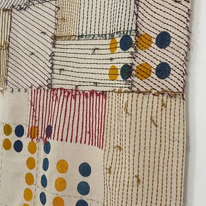 May include: A textile wall hanging featuring a patchwork of off-white fabric panels joined with visible stitching. The design includes sections with black, red, and gold vertical lines, alongside patterns of solid blue and gold circular dots.