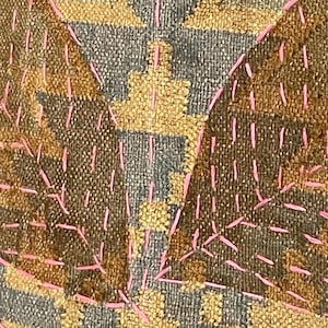 May include: Close-up of a textile with a geometric pattern in shades of brown, grey, and yellow. The design is accented with pink stitching, creating a textured and visually interesting surface. The fabric appears to be woven.