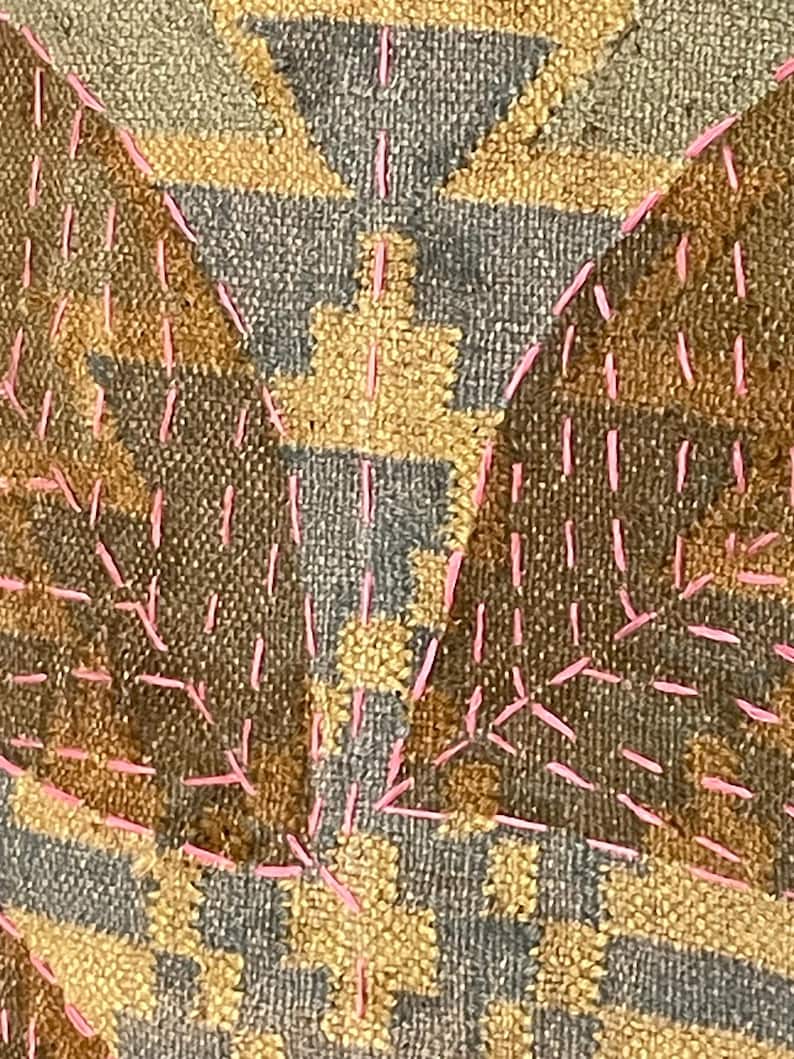 May include: Close-up of a textile with a geometric pattern in shades of brown, grey, and yellow. The design is accented with pink stitching, creating a textured and visually interesting surface. The fabric appears to be woven.