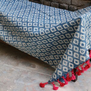 Peut inclure: Une couverture en coton bleu et blanc &agrave; motifs avec des glands rouges. La couverture est pli&eacute;e et drap&eacute;e sur un banc en bois.