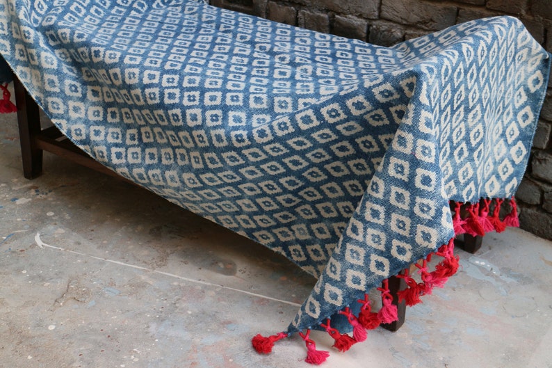 Peut inclure: Une couverture en coton bleu et blanc &agrave; motifs avec des glands rouges. La couverture est pli&eacute;e et drap&eacute;e sur un banc en bois.