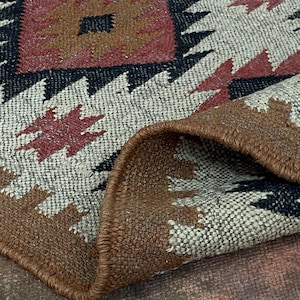 May include: Close-up of a woven rug with a geometric pattern. The rug features a beige background with red, black, and brown geometric shapes. The edge of the rug is folded over, revealing the underside.