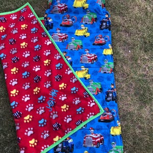 May include: A red and blue sleeping bag with a Paw Patrol theme. The sleeping bag has a red fleece blanket with paw prints and the name "JOHN" embroidered on it. The sleeping bag is made of a blue fabric with a repeating pattern of Paw Patrol characters and vehicles.